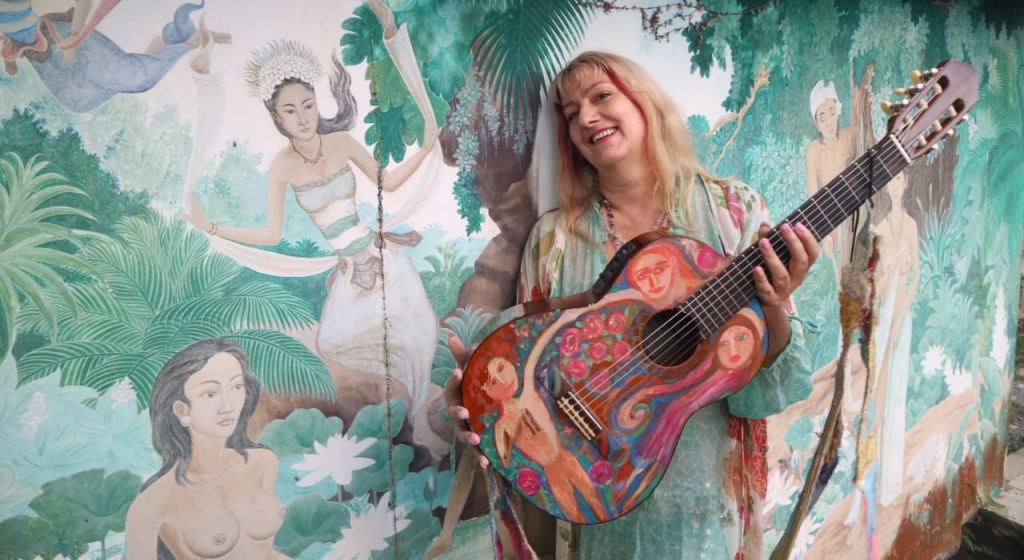 🎸 Jelila by the Pool of Light  
Against the shimmering mural by her swimming pool, Jelila cradles her hand‑painted guitar — each brushstroke alive with healing energy. The scene radiates artistry and serenity, a fusion of music, colour, and spirit that invites you to step into her world of transformation.